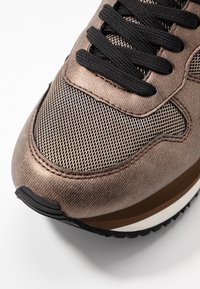 Bronze mesh and fabric sneaker with black laces, round toe, and layered sole featuring white and brown accents. Textured and breathable design.