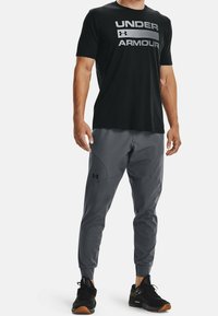 Muž v černém tričku Under Armour, šedých joggingových kalhotách s logem a černých sportovních botách stojí na bílém pozadí.