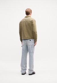 Man standing facing away wearing an olive green jacket, light blue loose jeans with a side pocket, and black shoes against a plain white background.