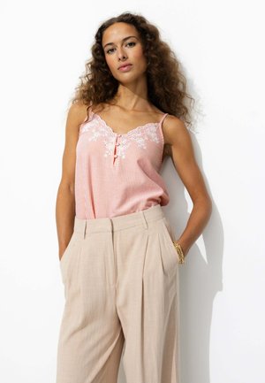 Camisole rayé rose clair avec des détails en dentelle blanche, présentant un détail de bouton à l'avant. Associée à un pantalon large plissé beige.