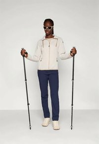 Gaišas krāsas zip-up jaka ar kontrastējošiem rāvējslēdzējiem, tumši zila bikses un krēmkrāsas apavi. Siluets tur melnās kalnu nūjas un valkā saulesbrilles.