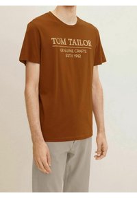 Hnědý bavlněný tričko s kulatým výstřihem, s krémovým logem "TOM TAILOR" a potiskem "GENUINE CRAFTS EST // 1962" na přední straně.