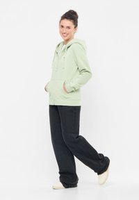 Felpa zip con cappuccio verde chiaro, dotata di lacci, tasche frontali e orlo a coste, abbinata a pantaloni neri larghi e scarpe di colore chiaro.