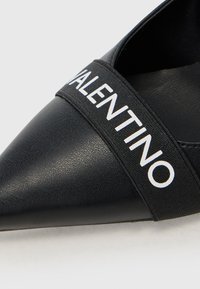 Sorte lær pointe flade sko har en elastisk rem med "VALENTINO" i hvid skrift. Designet er strømlinet med en glat tekstur.