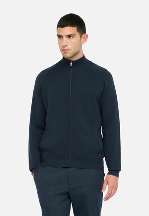 Marine blauwe zip-up jas met hoge kraag, geribde manchetten en zijzakken. Gemaakt van soepe stof, met een kleine rode accent bij de zoom.