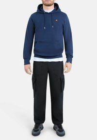 Hombre vestido con sudadera navy azul con logo, camiseta blanca debajo, pantalones cargo negros y zapatillas negras y grises, de pie contra un fondo blanco.