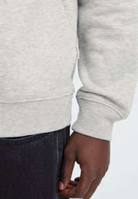 Sweatshirt gri cu manșete ribbate, etichetă mică de marcă pe lateral și o croială lejeră. Purtați cu pantaloni din denim închis.