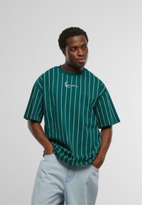 Zaļš svītrains īso piedurkņu T-krekls ar baltām vertikālām līnijām un apaļu kakla izgriezumu. Ieteikts izšūts logotips uz priekšpuses.