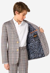 Abito a quadri con tonalità marroni, crema e grigie, accompagnato da una camicia bianca. L'interno presenta uno sfondo blu scuro con motivi grafici bianchi e un accento arancione.