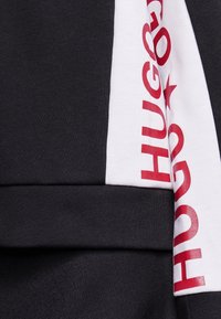 Czarna bluza z materiału z białą wstawką z czerwoną napisami "HUGO". Gładka tekstura, nowoczesny design oraz kontrastowe detale kolorystyczne.