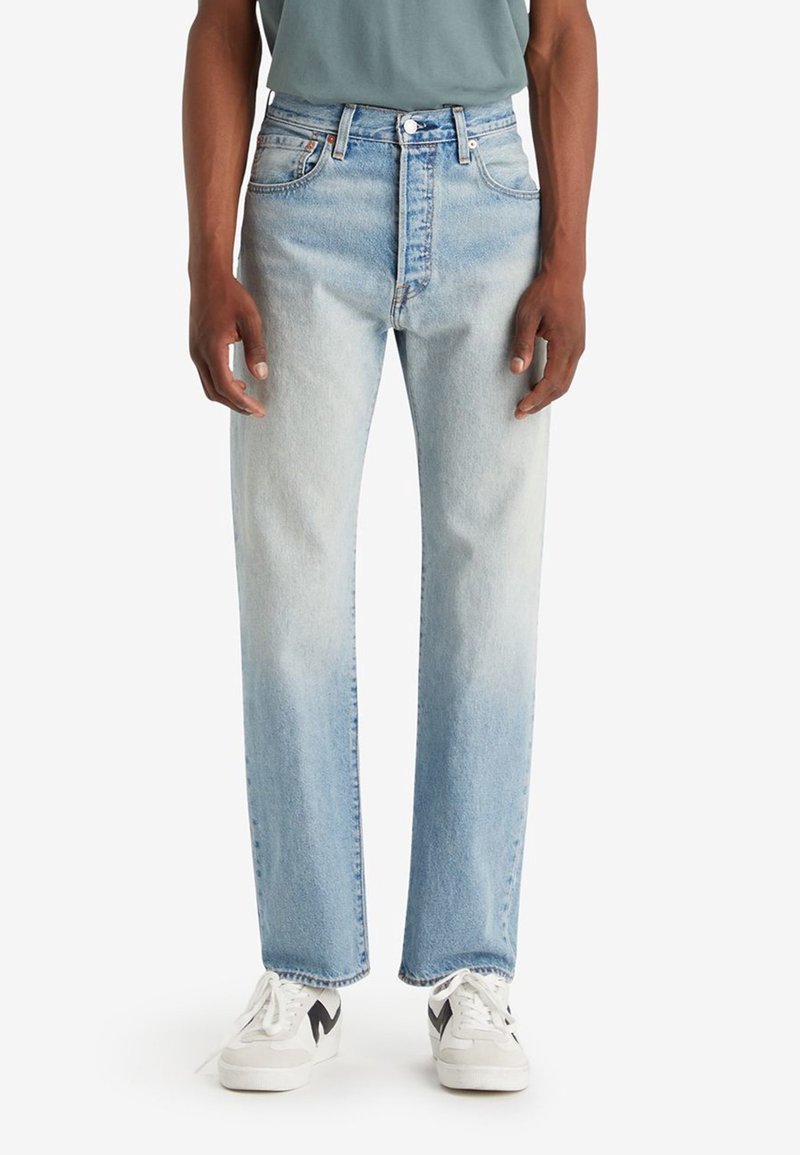 Homme portant un jean droit bleu clair et des baskets blanches avec des touches noires, debout devant un fond blanc.