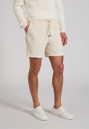 Man draagt beige shorts met trekkoord, witte sneakers en een crèmekleurige geribde trui met lange mouwen, staat met één hand in de zak.