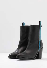 Bottes basses en cuir noir avec un bout pointu et un talon haut. Présentent un accent en zigzag contrasté bleu et argenté sur le côté.
