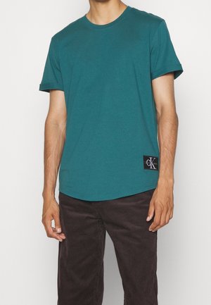 Mężczyzna w turkusowej koszulce z krótkim rękawem z naszywką z logo Calvin Klein i ciemnobrązowych spodniach sztruksowych, stojący na jasnym, jednolitym tle.