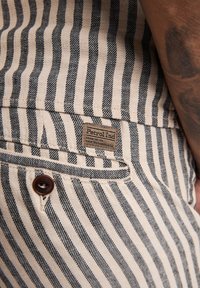 Gestreepte, beige stof met zwarte lijnen, voorzien van een bruine knop, een patchlabel "Petrol Ind" en een detail van een zijzak.