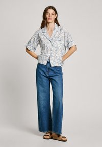 Kortermet skjorte i hvit med blå blomsterdesign, kombinert med høyhårede blå jeans og brune sandaler, stående mot en lys bakgrunn.