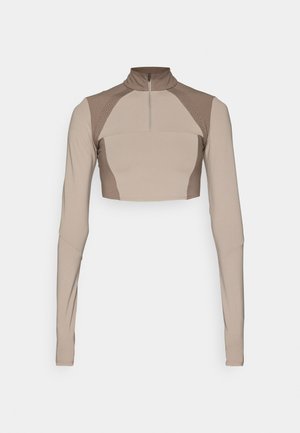 Haut beige coupé, à manches longues avec un col montant, fermeture éclair quart et détails de design texturé aux épaules. Fabriqué en tissu lisse et extensible.