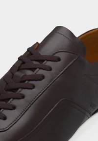 Baskets en cuir marron foncé avec des lacets assortis, des coutures nettes et une doublure intérieure couleur beige, présentées en gros plan.