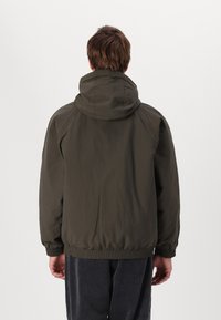 Veste verte, légère, à capuche avec des poignets et un ourlet élastiques. Présente une texture lisse et un design épuré et minimaliste.