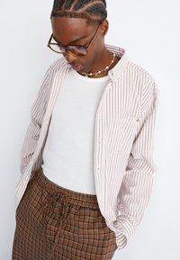 Mann mit geflochtenem Haar, der eine Brille aus Schildpatt, ein weißes Tanktop, ein lockeres gestreiftes Hemd und eine braun karierte Hose trägt und nach unten schaut.