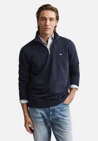 Maglione blu navy con zip, polsini e orlo a coste, indossato sopra una camicia a righe chiara con colletto, abbinato a jeans azzurri.