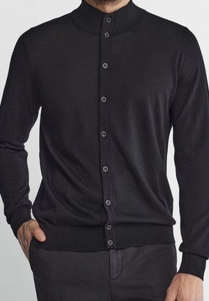 Homme portant un cardigan noir cintré à boutons, avec des poignets et un ourlet côtelés, une main dans la poche, sur un fond neutre.