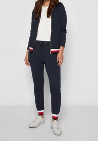 Sudadera con cremallera azul marino y cordones, camiseta interior blanca y pantalones azul marino con puños a rayas rojas y blancas, combinados con zapatillas blancas.