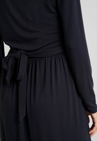 Mörk marinblå klänning med långa ärmar, en rynkad midja med en knytbanedetalj och en mjuk, slät tygkvalitet.