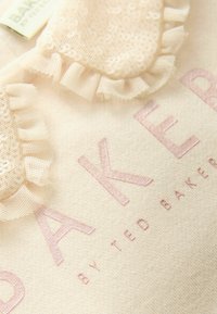 Bež kangas koos roosa tikandiga tekstiga "BAKER", millel on pitsilaadne rümpakujuline aktsent. Pehme tekstuur ja õrnad detailid.