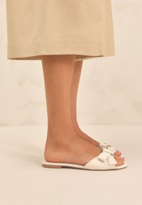 Sandales à glissière en cuir blanc avec un grand nœud, une semelle plate et un design à bout ouvert. Des jambes sont posées sur un fond clair.