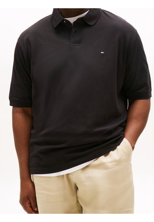 Mężczyzna w czarnej koszulce polo z krótkim rękawem i małym logo na piersi oraz beżowych spodniach, z jedną ręką w kieszeni.