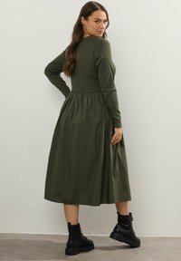 Kobieta w oliwkowej, długiej sukience midi z długim rękawem i czarnych, sznurowanych butach, stojąca z jedną ręką na biodrze, spoglądająca przez ramię.