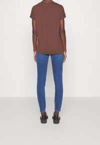 Persona vistiendo una camiseta de manga corta marrón, jeans ajustados azules y zapatos negros, de pie frente a una pared blanca sencilla.