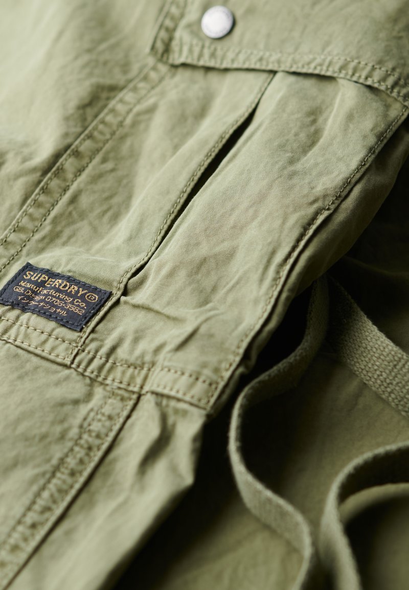 Olijfgroen stof met een gekreukeld textuur, voorzien van een voorzak en een marineblauwe label met "Superdry." Zilveren drukknoopsluiting aan de bovenkant.