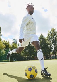 Atleta vestido con una camiseta y pantalones cortos blancos con detalles en negro, chutando un balón de fútbol amarillo y azul en un campo verde bajo un cielo nublado.