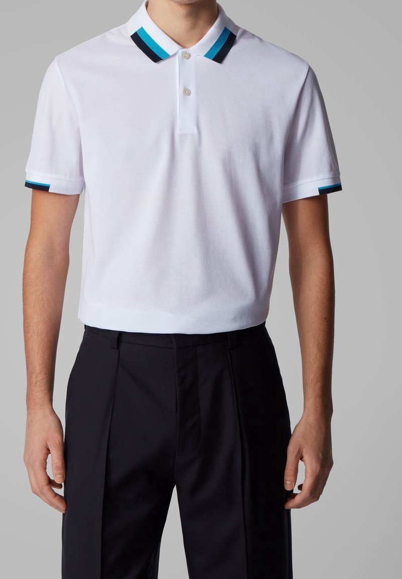 Polo shirt blanc avec un col à deux boutons, présentant des détails en rayures bleues et noires sur le col et les manches, associé à un pantalon noir.