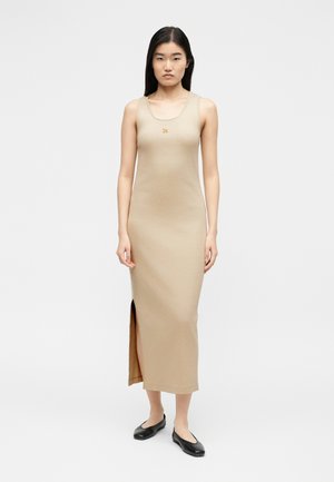 Γυναίκα στέκεται φορώντας μια μπεζ midi φόρεμα χωρίς μανίκια με πλαϊνή σχισμή και μικρό κεντημένο τίγρη στο στήθος, συνδυασμένο με μαύρα φλατ παπούτσια.