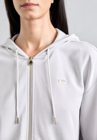 Sweatshirt cinza claro com fecho éclair e textura suave, apresentando um zíper dourado, cordões brancos e um pequeno logo "EA7" no peito.
