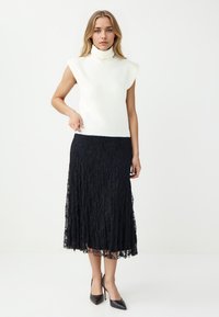 Hellbeige ärmelloses Rollkragenoberteil, kombiniert mit einem schwarzen Spitzen-Midirock; das Rockdesign hat ein strukturiertes Muster und eine ausgestellt Silhouette.