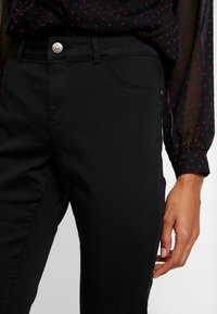 Černé džíny z denimu s přiléhavým střihem, s kovovým zapínáním na knoflík, bočními kapsami a diskrétními švy. Měkký bavlněný materiál.