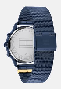 Tamsiai mėlynas nerūdijančio plieno laikrodis su tinkleliu, turi chronografo dizainą ir poliruotą nugarėlę su įraiža "TOMMY HILFIGER".