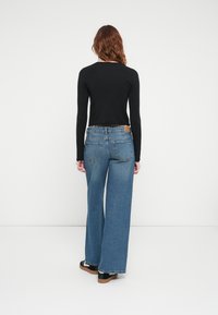 Bredde ben blå denimjeans med høy midje, med fem lommer. Svart langermet topp delvis synlig bak.