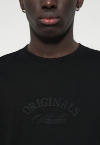 Camiseta de algodón negra con un logotipo bordado tonal "ORIGINALS Studio". El diseño incluye un cuello redondo clásico y una textura suave.