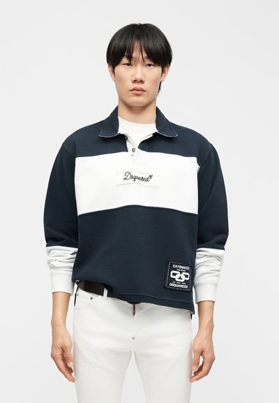 Νεαρός άνδρας με ίσια μαύρα μαλλιά, φορώντας μπλε ναυτικό και λευκό μακρυμάνικο ράγκμπι μπλουζάκι και λευκό παντελόνι, στέκεται μπροστά σε απλό φόντο.