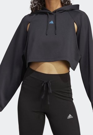 Kobieta ubrana w czarną krótka bluzę z wycięciami na ramionach oraz czarne legginsy Adidas ze sznurkiem w pasie.