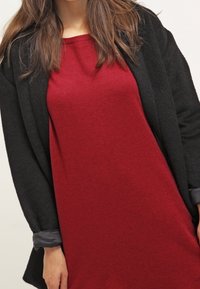 Cardigan noir à ouverture frontale sur une robe pull rouge. Le cardigan a une texture douce, tandis que la robe est en tissu lisse et chaud.