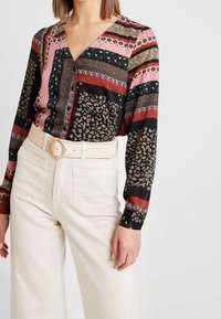 Blusa a maniche lunghe con scollatura a V, caratterizzata da un mix di fantasie in nero, rosa, verde e stampa leopardata, abbinata a pantaloni a vita alta di colore crema.