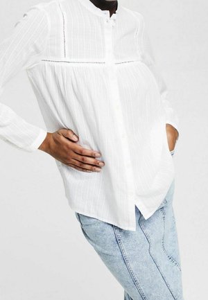 Person iført en løs hvid tekstureret skjorte med knapper og blonder samt lyseblå jeans, poserer med den ene hånd i lommen.