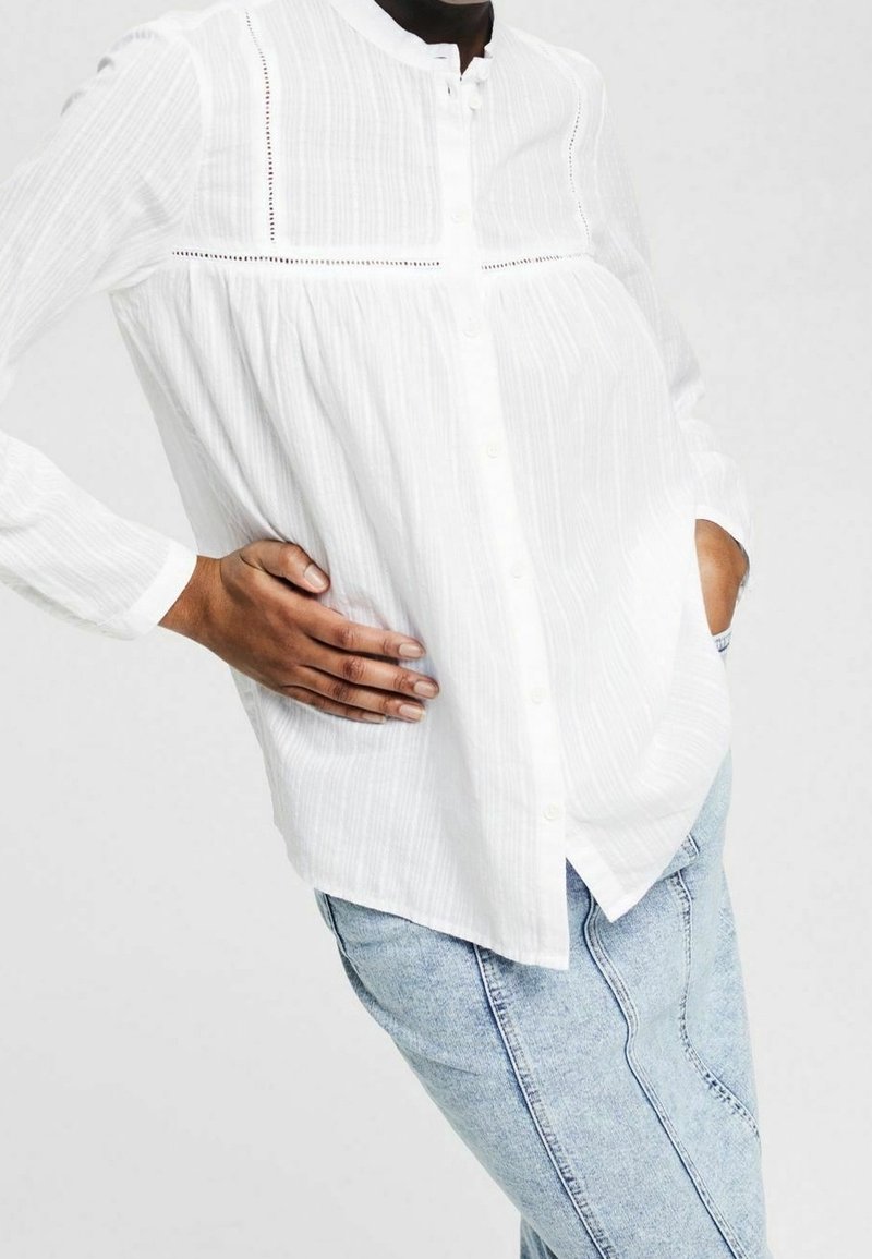Personne portant un chemisier blanc texturé et ample à boutons avec bordure en dentelle, et un jean bleu clair, posant avec une main dans la poche.