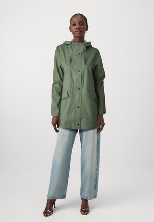 Veste de pluie verte imperméable avec capuche, manches longues, boutons-pression et deux poches à l'avant, associée à un jean large bleu clair.